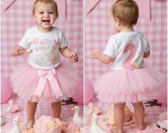 Può includere: Un bambino indossa una maglietta bianca con la scritta "silly goose" e una gonna tutu rosa. Intorno, una torta rosa, oche giocattolo e palloncini rosa. Lo sfondo è un motivo a quadretti rosa.