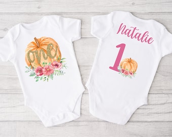 Può includere: Due body bianchi con un design a forma di zucca e fiori. Un body dice "one" in glitter dorati su una zucca e l'altro body dice "Natalie 1" in rosa.