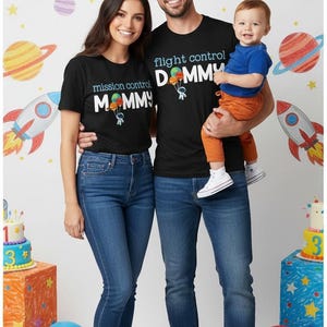 May include: A family of three wearing matching black t-shirts with space-themed graphics and text. The shirts say "Mission Control Mommy" and "Flight Control Daddy." The background features a space-themed banner and decorations.