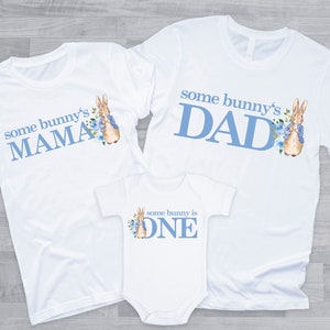 Puede incluir: Tres camisetas blancas con texto azul y una ilustraci&oacute;n de un conejito. Las camisetas dicen "some bunny's MAMA", "some bunny's DAD" y "some bunny is ONE".