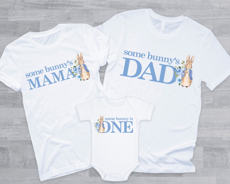 Puede incluir: Tres camisetas blancas con texto azul y una ilustraci&oacute;n de un conejito. Las camisetas dicen "some bunny's MAMA", "some bunny's DAD" y "some bunny is ONE".