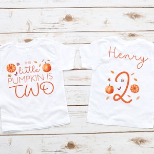 May include: Two white t-shirts with a fall theme. One shirt says "This little pumpkin is two" with a pumpkin and leaves. The other shirt says "Henry" with a number two and pumpkins.