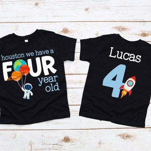 May include: Black t-shirt with a white astronaut holding balloons with planets and the text "houston we have a FOUR year old". The back of the shirt has a blue number 4 with a rocket and the name "Lucas" in white.