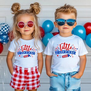 Puede incluir: Dos niños con camisetas blancas con el texto "Little Firecracker" impreso en ellas. La niña lleva pantalones cortos a cuadros rojos y blancos y gafas de sol. El niño lleva pantalones vaqueros azules y gafas de sol.