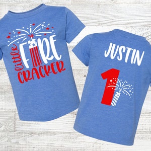 Puede incluir: Una camiseta azul con un diseño de fuegos artificiales rojo y blanco y el texto "little firecracker". La parte posterior de la camiseta tiene un número uno rojo con un diseño de fuegos artificiales y el nombre "Justin".