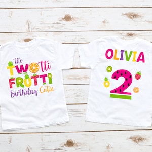 May include: Two white t-shirts with a fruit theme. The front of one shirt says "the twotti frutti birthday cutie" with a pineapple, orange, kiwi, and strawberry. The back of the other shirt says "OLIVIA" in rainbow colors and has a watermelon number 2 with a pineapple, kiwi, and strawberry.