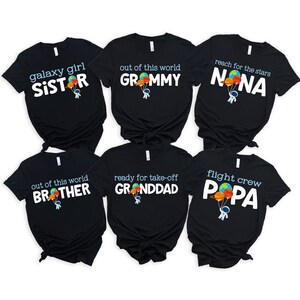 May include: Six black t-shirts with white text and a cartoon astronaut holding balloons. The shirts read: "galaxy girl SISTER", "out of this world GROMMY", "reach for the stars NONA", "out of this world BROTHER", "ready for take-off GRANDDAD", and "flight crew PAPA".