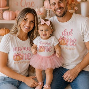May include: Three white t-shirts and a white baby bodysuit featuring pink pumpkin graphics with plaid bows and text reading mama of one, dad of one, and this little pumpkin is one. The baby wears a pink tulle skirt, a pink bow headband, and pink shoes.
