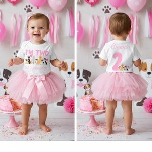 Op de afbeelding: Een peuter in een wit T-shirt en een roze tutu-rok. Het shirt zegt "I'M TWO let's pawty!" met hondenillustraties. De achterkant van het shirt heeft de naam "Gianna" en het nummer "2". Een roze strik siert de tutu.