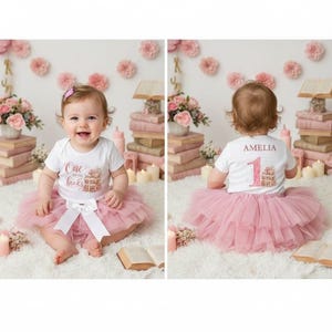 May include: A baby wearing a white onesie with the text "One books" and a pink tutu skirt. The onesie has a teddy bear and book design. The baby is sitting on a white fluffy rug. The background includes books, flowers, and candles.
