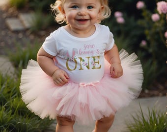 Puede incluir: Una niña pequeña con una camiseta blanca con el texto "Some bunny is ONE" y un tutú rosa. La niña también lleva una diadema con orejas de conejo rosas y zapatillas blancas. La niña sonríe y está de pie en un camino de piedra.