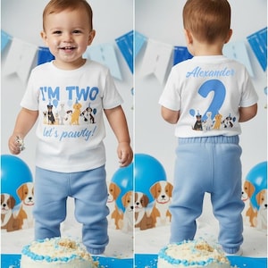 Op de afbeelding: Een peuter in een wit T-shirt en een blauwe broek viert zijn tweede verjaardag. Op de voorkant van het shirt staat "I'M TWO" met een hondenafbeelding en "let's pawty!". De achterkant van het shirt zegt "Alexander" en heeft een nummer 2. Een blauwe en witte taart staat vooraan.