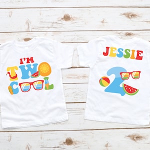 以下が含まれることがあります： カラフルな夏のグラフィックが施された2枚の白いTシャツ。1枚はフラミンゴ、サングラス、フルーツと共に「I'M TWO COOL」と書かれています。もう1枚は「JESSIE」と書かれており、サングラスとスイカのスライスが付いた数字の2が表示されています。