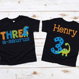 May include: Black toddler t-shirts with dinosaur graphics. One shirt says "THREE a-saurus" with a blue dinosaur. The other shirt says "Henry 3" with a green and yellow dinosaur. The text and graphics are in orange, blue, and green.