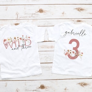 Może przedstawiać: Dwie białe koszulki dla maluchów. Jedna koszulka ma napis "WILD and three" z kwiatowymi akcentami. Druga koszulka ma imię "gabrielle" i cyfrę "3" z kwiatowymi akcentami. Obie koszulki znajdują się na jasnym drewnianym tle.