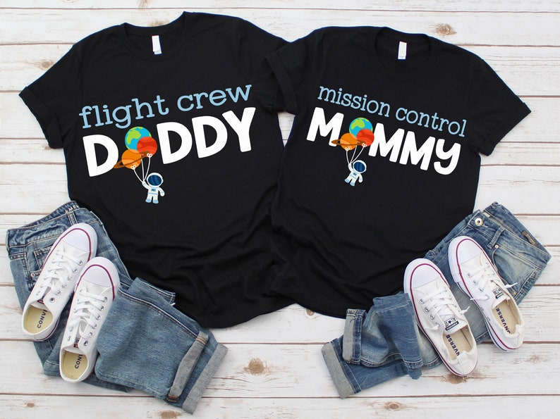 May include: Two black t-shirts with white text. One says "flight crew DADDY" and the other says "mission control MOMMY". Each shirt has a graphic of an astronaut holding balloons. Blue jeans and white trainers are also in the image.