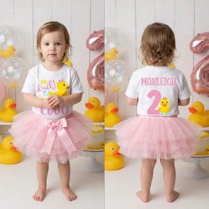 May include: A toddler wearing a white t-shirt with a yellow duck graphic and the text "oh duck! two". She is wearing a pink tutu skirt with a bow. The background includes rubber duckies and balloons.