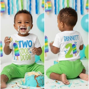 May include: A toddler wearing a white t-shirt with the text "TWO COOL DUDE" and a popsicle graphic, paired with green pants. The child is covered in cake frosting, with a blue cake in front. The background includes balloons and streamers.