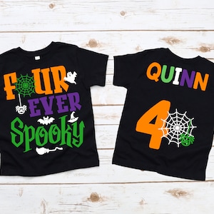 Op de afbeelding: Twee zwarte T-shirts met Halloween-thema's. Het voorste shirt zegt "Four Ever Spooky" met een spinnenweb, een spin, een vleermuis en een heksenhoed. Het achterste shirt zegt "Quinn" in regenboogkleuren en heeft een groot oranje nummer 4 met een spinnenweb en een spin.