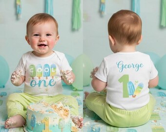 May include: A baby wearing a white t-shirt with the words "Cool One" and a matching cake. The baby is also wearing light green pants. The cake is decorated with blue frosting and a number 1.