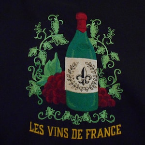 May include: Embroidered design of a green wine bottle with a white label featuring a fleur-de-lis and the text "LES VINS DE FRANCE" in gold lettering. The bottle is surrounded by green vines and red grapes.