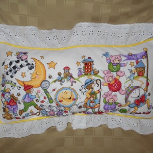May include: A white pillow with a yellow border and a white lace trim. The pillow features a colorful embroidered design of animals and characters, including a cow jumping over the moon, a pig wearing a number 2, and a mouse playing a drum. The design also includes stars, a house, and a tree.