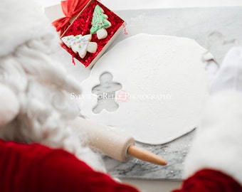 May include: Santa Claus in a red suit and white beard is making gingerbread cookies. He is using a gingerbread man cookie cutter and a rolling pin to roll out the dough. The dough is on a white surface and there are two decorated Christmas tree cookies in a box next to him.