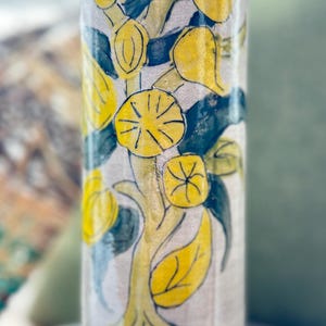 May include: A tall, cylindrical ceramic vase with a hand-painted lemon tree design. The vase features yellow lemons, green leaves, and a yellow trunk against a beige background. The vase is filled with dried stems.