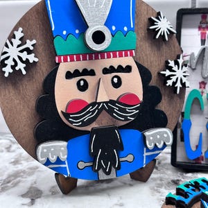 May include: A wooden Christmas ornament featuring a nutcracker soldier with a blue hat and coat, black hair and mustache, and a tan face. The ornament is round with snowflakes and is on a small wooden stand.
