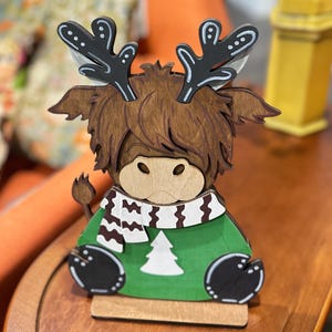 May include: Wooden Highland cow figurine with a green sweater, white and black scarf, and black antlers. The cow has a light brown face and a white Christmas tree design on the sweater. The figurine is sitting on a wooden base.