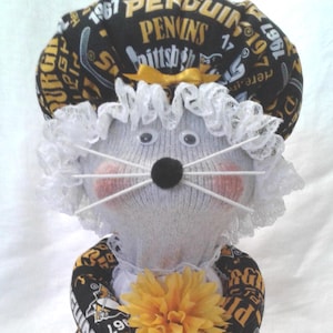 May include: A handmade sock doll dressed in a Pittsburgh Penguins-themed outfit. The doll has a gray body, a black nose, and white whiskers. It wears a hat and apron with the team's logo and colors, and holds a yellow flower.