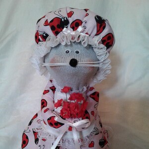 May include: A handmade sock doll dressed in a pink dress with a ladybug print and a white lace trim. The doll is wearing a pink and white ladybug print hat with a white lace trim. The doll is holding a bouquet of red flowers.