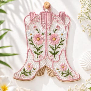 May include: A decorative wall hanging shaped like a pair of cowboy boots, crafted from white lace with a hexagonal mesh pattern. The boots feature embroidered pink and white daisy flowers with green stems and tan heels, suspended by a small wooden peg.
