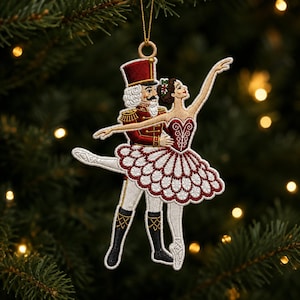 May include: A hanging Christmas tree ornament featuring an embroidered nutcracker soldier in a red uniform and a ballerina in a white and red tutu. The nutcracker wears a tall red hat and black boots, while the ballerina has her arm raised.