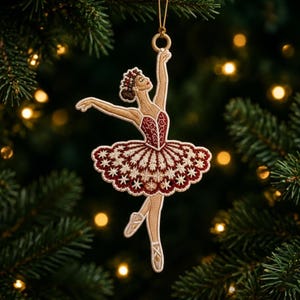 May include: A hanging ornament shaped like a ballerina in a pose with one arm raised. The figure features intricate embroidery in shades of deep red, cream, and gold, with a detailed tutu adorned with star-like patterns and a gold hanging loop.