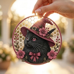 May include: A circular embroidery hoop art piece featuring a black cat wearing a maroon witch hat with a buckle and pink flowers. The cat has pink eyes and a matching pink bow tie. The background is a white honeycomb mesh with floral accents.