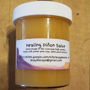 Puede incluir: Un tarro de Healing Piñon Salve con tapa blanca. La etiqueta dice "Healing Piñon Salve" y detalla los ingredientes y la información del fabricante. La pomada del interior es de color ámbar claro. El tarro es de cristal transparente.