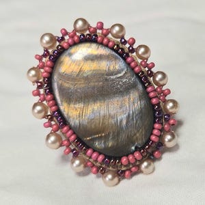 May include: An oval ring featuring a central tiger's eye gemstone with brown and gold chatoyancy. The stone is surrounded by a border of dark purple beads, followed by a ring of matte pink beads, and finished with an outer edge of cream pearls.