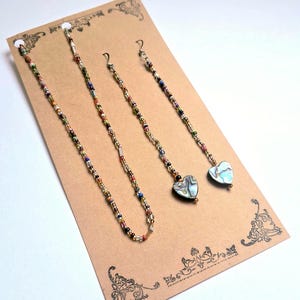 Pode incluir: Um colar e dois pares de brincos exibidos em um cartão marrom. As joias apresentam contas coloridas e pingentes em forma de coração de concha de abalone. O colar e os brincos são enfiados em um fio fino de cor prateada.