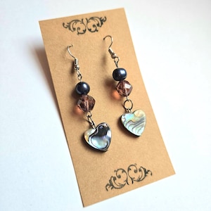 Pode incluir: Brincos pendurados com ganchos prateados. Cada brinco apresenta uma conta azul escura, uma conta castanha facetada e um pendente em forma de coração de concha de abalone. Apresentados num cartão castanho com desenhos decorativos nos cantos.