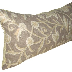 May include: A long, rectangular pillow with a beige linen fabric cover. The pillow is embroidered with a floral pattern in a lighter beige color.