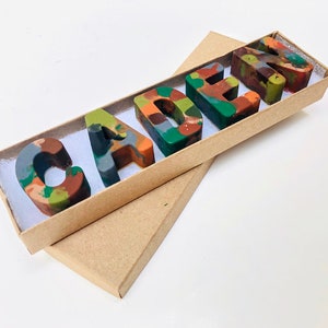 May include: A brown cardboard box with a white interior, containing a set of five colorful, multi-colored crayon letters spelling out the name "CADEN".