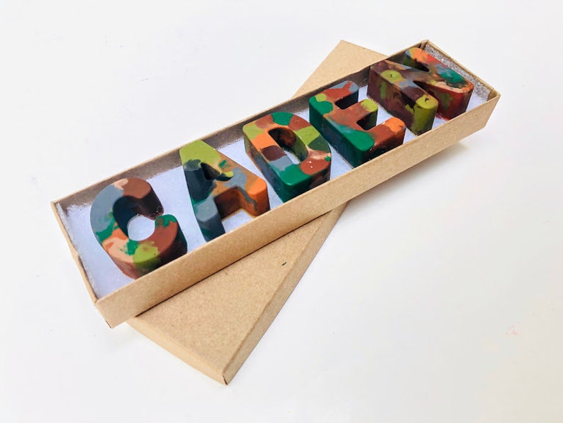 May include: A brown cardboard box with a white interior, containing a set of five colorful, multi-colored crayon letters spelling out the name "CADEN".