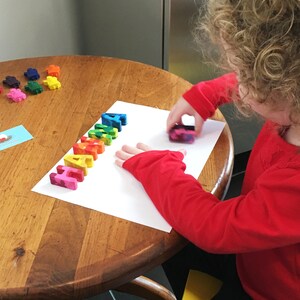 May include: A child uses colorful letter shaped crayons to spell the word "HAHA" on a white sheet of paper.