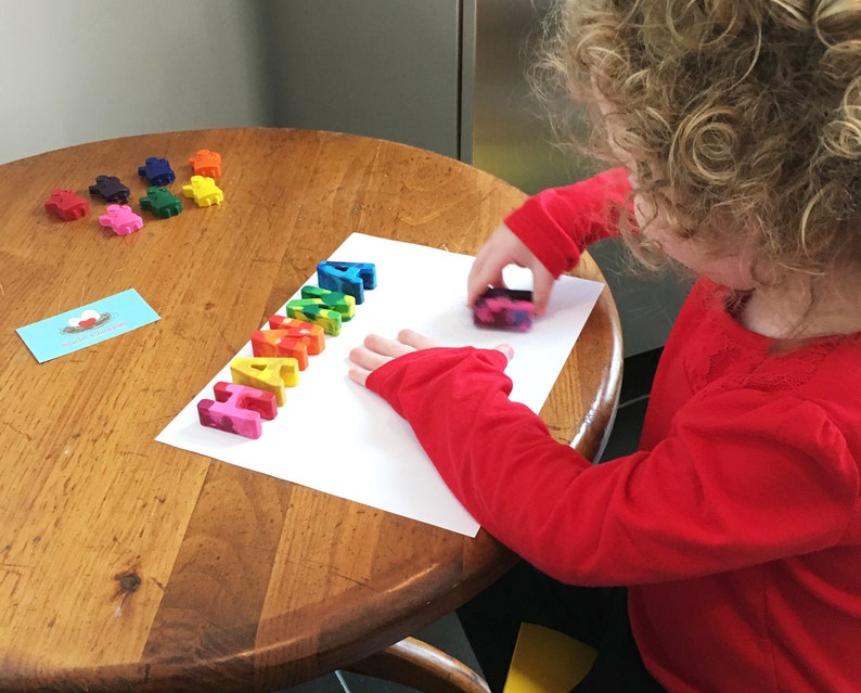 May include: A child uses colorful letter shaped crayons to spell the word "HAHA" on a white sheet of paper.