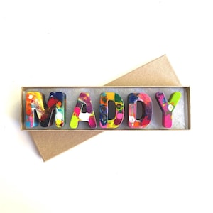 May include: A set of colorful crayon letters spelling out the name "MADDY" in a brown cardboard box.