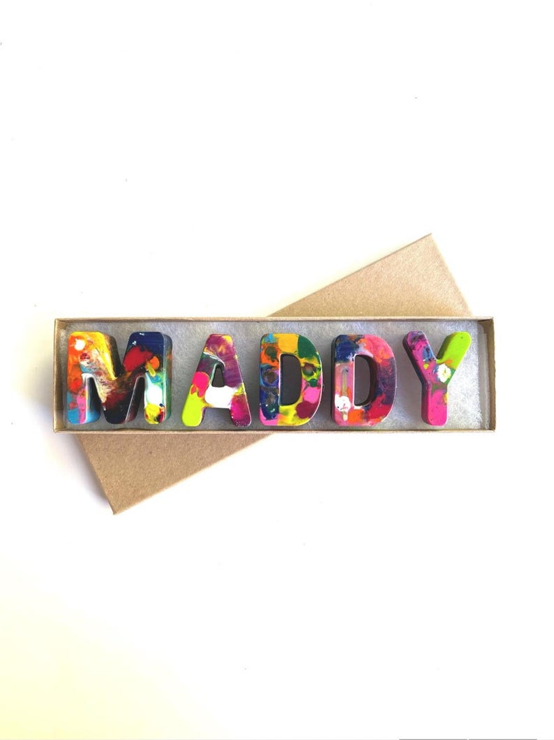 May include: A set of colorful crayon letters spelling out the name "MADDY" in a brown cardboard box.