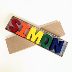 May include: A set of colorful crayon letters spelling out the name "SIMON" in a brown cardboard box.