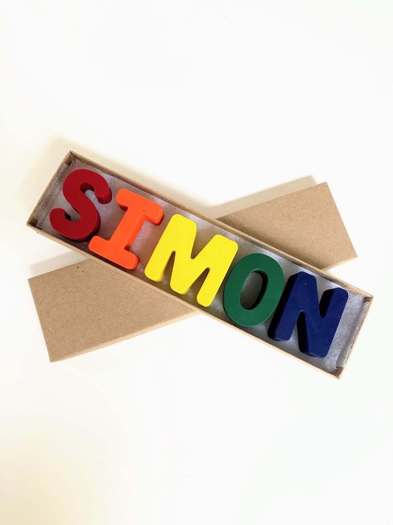 May include: A set of colorful crayon letters spelling out the name "SIMON" in a brown cardboard box.
