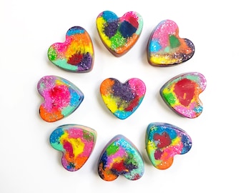May include: Eight heart-shaped crayons in various colors, including red, blue, green, yellow, and purple. The crayons have a glittery finish and are arranged in a circle on a white background.